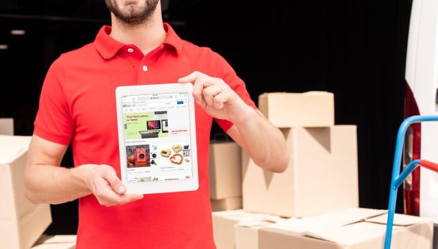 Smiling mover in red shirt holding a tablet in front of moving boxes, representing how moving companies can improve visibility with local SEO services.