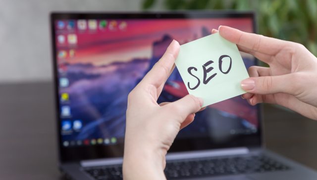 Hands holding a sticky note with "SEO" written on it in front of a laptop screen.